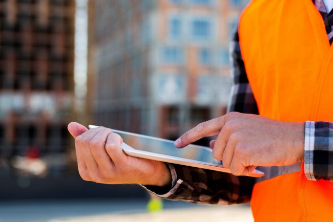 Closeup of man in safety vest holding tablet illustrates blog "The Pandemic and the Rising Costs of Building"
